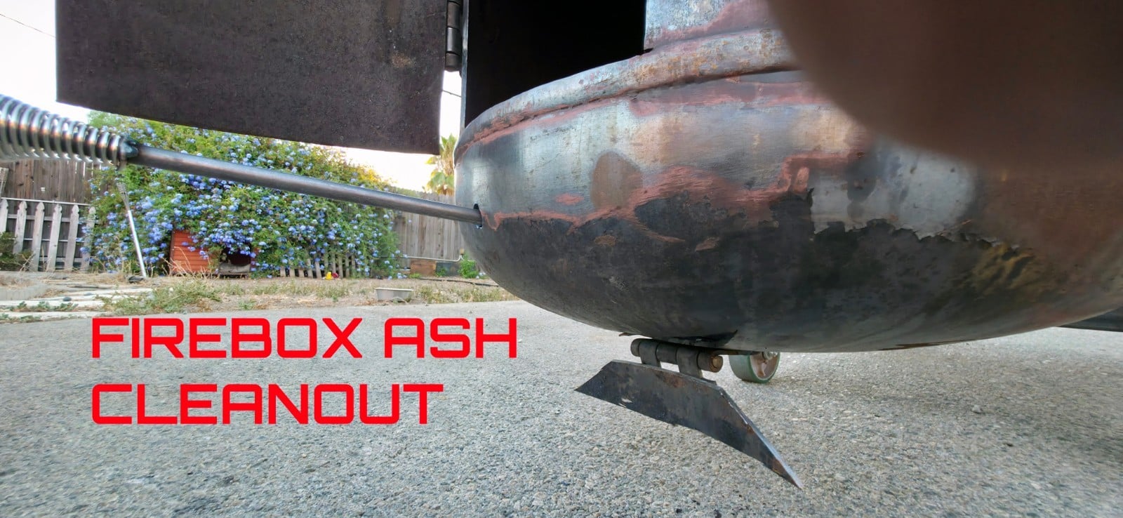Firebox ash cleanout at the base of the vertical tower being raked out with a long handle while the smoker sits on its casters
